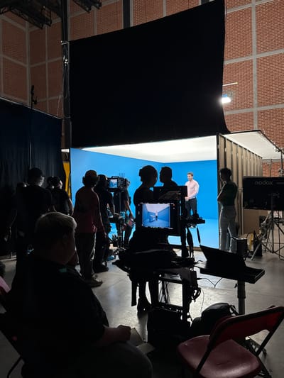 The image shows a film set with a blue screen backdrop in what appears to be a large studio or warehouse with a brick ceiling. Several people are present, some operating camera equipment and others watching the monitor or observing the scene. One individual is standing in front of the blue screen. There are various pieces of film equipment, including cameras, lighting, and monitors. A 'Godox' branded electronic device is visible. The scene is dimly lit, but the blue screen and the subject in front of it are well-lit. The location is likely an industrial or commercial area, potentially within the city of Tlalnepantla, Mexico.
