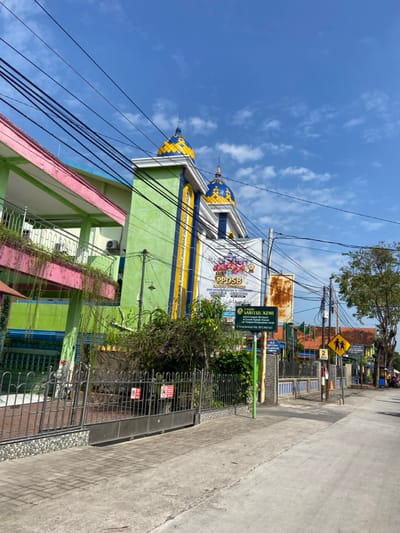 The image shows a street scene in the Kecamatan Tulangan area of Indonesia, located at coordinates -7.483, 112.65.  A two-story building with a green and pink facade stands on the left side of the street. The building has a distinctive feature: two domed structures on the roof, painted in a yellow and blue pattern. A sign on the building reads "PPDSB", likely referring to a local educational institution. The street is paved and empty, with a few power lines running overhead. A black metal fence separates the sidewalk from the street.  A green bush, and a few smaller plants grow along the front of the building. A signpost with the inscription "PAUD SABITUL AZMI" stands on the right side of the fence.  A  "children crossing" sign is visible in the background.  The sky is blue with white clouds.