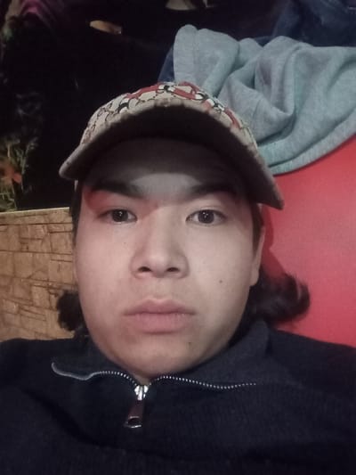 The image displays a close-up portrait of a male individual, observed in Takhtakuprik, Uzbekistan. The subject, appearing to be of East Asian or Central Asian descent, faces directly forward, looking into the camera with a neutral expression. He wears a beige-colored baseball cap with a patterned design, including red elements, its brim casting a shadow across his upper face. Dark hair is visible extending from beneath the cap on the sides. The subject is dressed in a dark blue or black collared sweater or jacket, featuring a silver zipper partially unzipped at the collarbone.
Behind the subject's right side, a red upholstered surface is visible, partially obscured by a light gray folded garment and an additional dark fabric item. To the subject's left, a textured wall with horizontal brown patterns is present. The background appears dark and indistinct, suggesting an indoor or covered setting, potentially during evening or night hours.