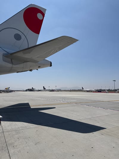 The image shows an airport tarmac in Tijuana, Mexico. The tail of an aircraft with a circular red and white design is in the foreground. Multiple other aircraft are visible in the background, parked along the taxiway. The concrete tarmac has lane markings. The shadow of the tail section stretches across the tarmac. The sky is clear and blue.