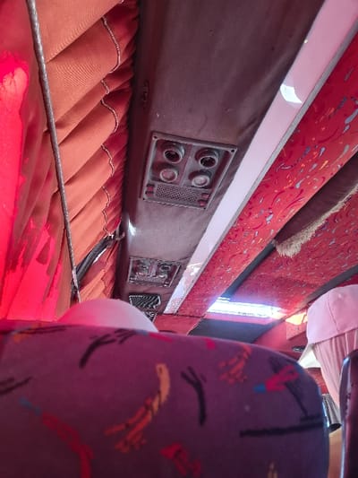 The image displays the interior of a passenger bus, potentially situated in Al-Kiswa, Syria, although the specific geographical location is not verifiable from the visual evidence alone. The vantage point is from a low angle, looking forward and upward within the vehicle.

The ceiling is composed of dark brown fabric paneling. Mounted centrally on this paneling is a rectangular fixture containing four circular, black air nozzles or vents arranged in a 2x2 grid, positioned above a rectangular grille. Further along the ceiling, a second, smaller, similar fixture is partially visible. A long, white, linear fluorescent light fixture is installed along the right side of the ceiling.

The upper sections of the bus's interior walls are covered with pleated reddish-orange fabric, serving as either curtains or decorative upholstery. A dark cable descends vertically on the far left side of the frame, adjacent to the fabric.

In the foreground, the upper portion of a bus seat is visible, upholstered in a dark purple fabric adorned with abstract, multi-colored patterns in shades of yellow, orange, red, and light blue. To the right, a cream or light-colored fabric headrest cover is present over another seat. At the upper distant end of the ceiling, a rectangular opening, consistent with a sunroof or skylight, reveals bright exterior light. No individuals or specific actions are depicted within the scene.