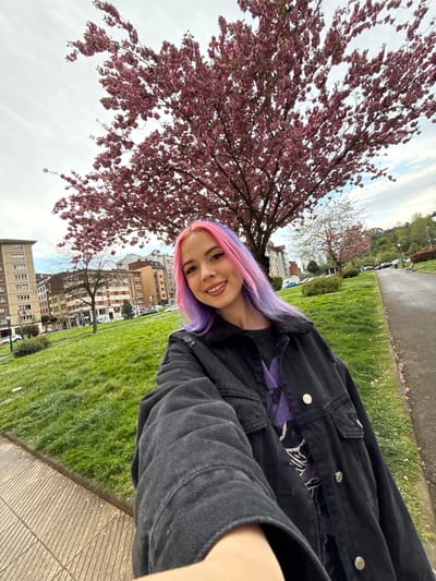 The image shows a young woman with pink and purple hair taking a selfie outdoors. She is wearing a black denim jacket over a graphic t-shirt. Behind her is a flowering cherry tree with pink blossoms. In the background are apartment buildings and a green lawn, suggesting a park or residential area. The location appears to be Oviedo/Uviéu, Spain, based on the architectural style of the buildings.