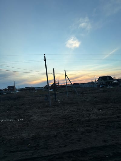 The image depicts a rural scene, likely on the outskirts of the city of Дулесово, Russia, given the presence of utility poles and sparse buildings against a backdrop of open land. The landscape is dominated by a field of dry, withered vegetation, suggesting a season like late autumn or early spring. Utility poles stand prominently in the foreground, with electrical lines stretching across the sky. A few houses are visible in the distance, their silhouettes outlined against the sunset. The sky is gradient of blues and oranges, characteristic of twilight.