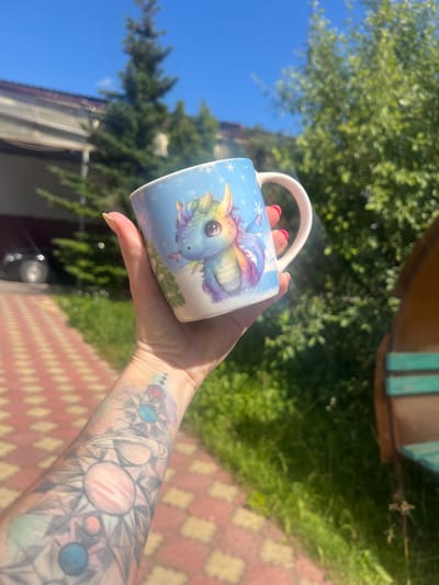 A person is holding a mug with a colorful dragon design on it. The mug is white with a light blue background and features a cartoon dragon with large eyes, purple scales, and rainbow-colored wings. Snowflakes are also visible on the mug's surface. The person's hand, with neatly manicured nails, is holding the mug. Their forearm is visible and adorned with a tattoo depicting celestial bodies, including planets and stars, in an abstract, geometric style.

The scene appears to be outdoors on a sunny day, indicated by the bright sunlight casting shadows and the clear blue sky. Lush green trees and foliage form the background, suggesting a natural or garden setting. A portion of a red and beige tiled walkway is also visible in the foreground, adding a touch of detail to the location. The overall impression is one of a relaxed, leisurely moment, possibly enjoying a drink outdoors. No text is discernible in the image. The location is identified as Krasnoe-na-Volge, Russia.