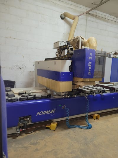 The image depicts a large blue and white CNC woodworking machine, labeled "FORMAT" and "profit 2.s", situated in a workshop environment. The machine is positioned against a textured, light-colored brick wall, which shows some signs of dirt and wear. The floor is a concrete surface, covered in a layer of dust and wood debris.
Several smaller, dark-colored mechanical components, possibly fixtures or clamps, are arranged in a scattered pattern on the blue surface of the machine in the foreground. A coiled blue pneumatic hose is connected to the machine and rests on the floor. A yellow foot pedal is also visible on the floor near the machine.
Above the machine, a large, flexible yellow duct connects to an exhaust system, suggesting dust extraction during operation. The machine itself features various mechanical components, including a track system with yellow and black elements, and a protective clear enclosure on the right side. In the background, stacks of what appear to be wooden planks or sheets are visible.
The overall impression is a functional industrial setting, likely a furniture manufacturing or woodworking shop. Although the specific location of Łomianki, Poland is mentioned, there are no explicit visual cues in the image to confirm this geographical detail. The scene is static, with no people or active operations immediately evident.