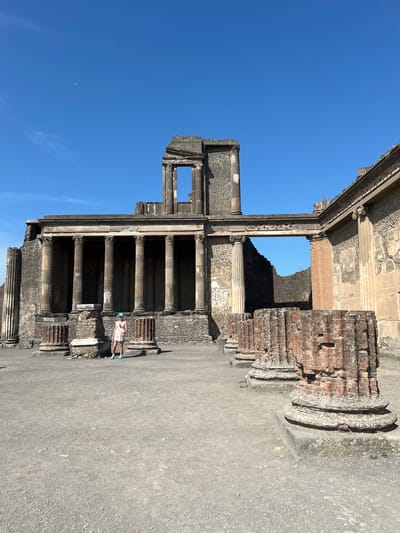 A person stands in the courtyard of an ancient Roman building, likely in the city of Pompeii, Italy. The person is wearing a light-colored dress, a blue baseball cap, and bright blue shoes, and is facing the camera. The building is in ruins, with stone columns and walls visible. The sky is clear and blue, with a faint moon visible. The ground is covered in gravel. The scene depicts a sunny day at an archaeological site.