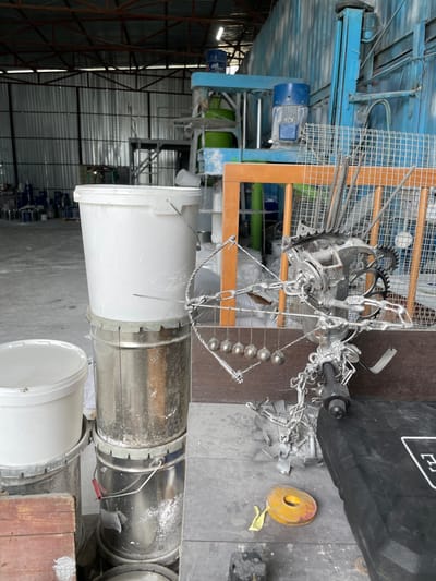 This is a medium shot, taken outdoors in what appears to be an industrial setting, possibly a workshop or manufacturing facility. The image is oriented vertically. In the foreground, a large, white plastic bucket sits next to a section of a metallic pipe, also appearing industrial. Behind these, more metallic pipes or conduits are visible. The background reveals a large building with a corrugated metal roof and a steel framework, suggesting a warehouse or factory.
To the left of the main subjects, a blue industrial machine with motors and wires is partially visible, hinting at an operational or recently used piece of equipment. Above and to the right of the bucket and pipe are components of a chain mechanism, including metal balls, chains, and what looks like a cogwheel. A wooden frame structure is also present, possibly part of a larger piece of machinery or scaffolding. A black tarp or bag with some white lettering is seen in the upper right quadrant of the image. A yellow circular object with a piece of yellow tape attached lies on the ground nearby. The overall impression is one of an active industrial environment, likely in Kazakhstan, given the provided location context. No people are visible in the frame. The lighting suggests it could be daytime, but the specific time of day or weather conditions are not discernible.