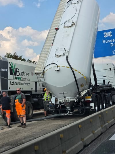 The image captures a scene on the B42 highway near Rüsselsheim am Main, Germany, where a large white cement mixer truck with a tilted silo is parked on the roadway. The truck bears the branding "Büttner Ökoservice Lohnunternehmen" and "SW 318 MC". Two men in work attire are present. One man, wearing orange overalls and a green shirt, stands facing the camera. Another man, in a black t-shirt and orange shorts, is partially visible to the left, leaning against the truck. A third person in a yellow vest is also visible near the rear of the cement mixer. The truck's silo is elevated, suggesting it is in the process of unloading or loading. The surrounding environment includes a concrete barrier separating the roadway and a blue highway sign indicating directions to Rüsselsheim and Darmstadt. The sky is partly cloudy.