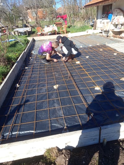 Two men are tying rebar together on a construction site, in the city of Stefanovo, Bulgaria. The site is a rectangular wooden frame with a black plastic liner. On top of the liner, a grid of rebar is being tied together by the men. One man is wearing a purple shirt, and the other is wearing a black hoodie. The men are squatting and using pliers to tie the rebar. The background shows a yard with tables, chairs, a house, and trees. The shadow of a person is visible in the foreground.
