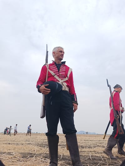 Two individuals in historical military uniforms are positioned in a dry, harvested agricultural field in Kottalapalli, India, under an overcast sky. The primary subject, a male with short grey hair, occupies the left foreground, standing with his head turned slightly to the viewer's left. He wears a red military jacket with white braiding and trim, dark trousers, and tall dark boots. A black shako-style hat is held in his left hand, and a long musket with an attached bayonet is slung over his right shoulder. A second male, also in a red military jacket, dark trousers, and boots, is partially visible in the mid-ground right, walking away from the camera. He wears a dark head covering and glasses, and carries a musket on his left shoulder. In the distant left background, several other individuals in civilian attire traverse the field. The ground consists of harvested light-brown stubble over dark soil. A low mountain range and sparse trees are discernible on the distant horizon.