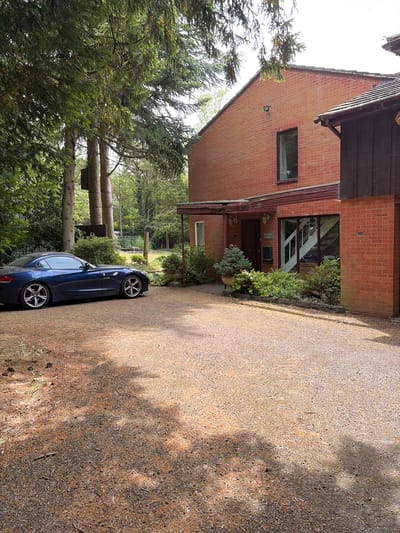 The image shows a two-story red brick house in a suburban setting, with a gravel driveway leading up to it. A blue car is parked on the driveway, partially obscured by trees. The house has a sign that reads "Victoria Lodge" near the entrance. The location is likely in North Hertfordshire, United Kingdom, judging by the style of the house and the surrounding landscape. There are no people visible in the image. The environment includes trees, foliage, and a well-maintained gravel driveway.