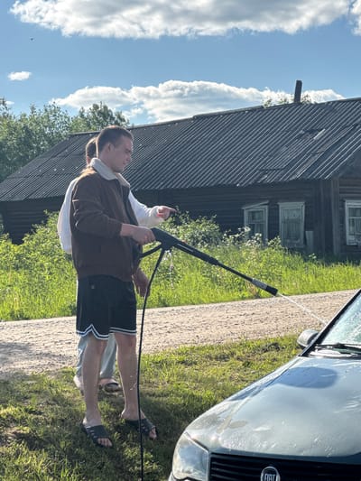 Two individuals are engaged in washing a light-colored car (identified as a Fiat by the visible badge on the grille) with a pressure washer outdoors. The primary subject, a man, stands in the foreground, holding the pressure washer lance and directing the water spray towards the car's front windshield. He is wearing a brown jacket with a white collar, black shorts with white athletic stripes, and dark sandals. Behind him, a second person, partially obscured, wears a white top and also appears to be assisting or observing the car washing.
The scene takes place on a sunny day, indicated by the bright blue sky with scattered white clouds and strong sunlight casting reflections on the wet car surface. The ground is a mix of a light-colored, possibly gravel or dirt, path and green grass. In the background, to the left, stands a weathered wooden building, likely a house or outbuilding, with visible windows and slatted siding, characteristic of a rural or village setting. This architecture and general environment align with the provided location context of Луптюг, Russia. The car's front is visible, covered in water from the washing.