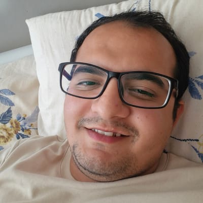 The image is a close-up, overhead selfie of a man wearing black-rimmed glasses, lying on a pillow with a floral pattern. The man has dark hair and a light complexion. He is wearing a light-colored t-shirt. His eyes are open, and he is smiling slightly. The background consists of a pillow and what appears to be a textured surface. There is no discernible information within the image to associate it with Küçükçekmece, Turkey.