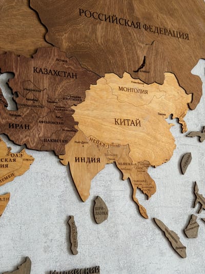 The image displays a close-up, high-angle shot of a wooden world map on a textured gray wall, possibly in Semiluki, Russia, given the Cyrillic inscriptions. The map is crafted from different shades of wood, with country outlines and names etched into the surface. Various countries and regions are depicted, including Russia, Kazakhstan, Mongolia, China, Iran, Afghanistan, Pakistan, India, Nepal, and parts of Southeast Asia. Some smaller island nations and peninsulas are also represented as separate wooden pieces. The map is intricately detailed, showcasing geographical features and labels in Russian. The lighting is even, highlighting the texture of the wood and the wall. There are no people or animals visible in the image.