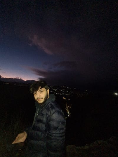 The image shows a single adult male standing on an elevated outdoor location overlooking an illuminated urban area at night. The location is Dummar Municipality, Syria. The male subject, positioned slightly left of center, has dark hair and a beard, and wears a dark blue or black puffy jacket over a dark hoodie. He is looking directly forward towards the camera. His left hand rests on an indistinct dark surface, possibly rock or ground, in the lower left foreground. The background features a sprawling cityscape or town illuminated by numerous artificial lights, indicating nighttime. The sky above is dark, transitioning from a deep twilight blue/purple near the horizon to a darker, almost black, overhead, with visible dark cloud formations. The immediate foreground consists of dark, indistinct terrain.
