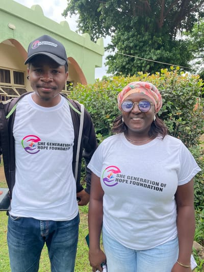 Two individuals, a man and a woman, stand outdoors in front of a light green building with arched doorways and windows. The man, on the left, wears a black baseball cap with a logo, a white t-shirt with the "SHE GENERATION OF HOPE FOUNDATION" logo, and a black zipped jacket over it. He is also wearing blue jeans. The woman, on the right, is wearing a headscarf, round glasses, earrings, a necklace, a white t-shirt with the same "SHE GENERATION OF HOPE FOUNDATION" logo, and light blue jeans. They are both smiling at the camera. Lush green foliage and a large tree with hanging roots are visible behind them. The ground is covered in grass. The building appears to be in a residential or institutional area within Unguwar Shanu, Nigeria.