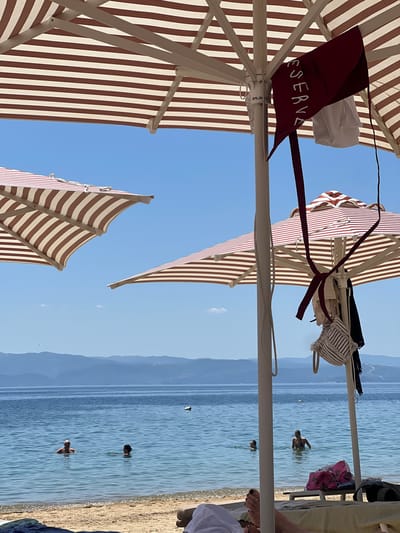 This image captures a sun-drenched beach scene in Troulos, Greece, with clear blue skies and calm, azure waters. In the foreground, striped beach umbrellas, predominantly in shades of brown and white, provide shade. One umbrella features a maroon accessory with white lettering that reads "RESERVE," and several garments, including a bikini top and what appears to be a white towel, are casually hung from its pole. Another umbrella is visible in the middle ground, also striped, and a third is angled in the upper left of the frame.

The sea is populated by several individuals. Two men are visible in the water, one closer to the shore and another further out. A woman is also in the water, and another person's head and shoulders are just above the surface. A yellow buoy is floating in the distance. The shoreline is sandy, and lounge chairs with towels are visible on the beach. In the far distance, a range of hills or mountains forms the horizon. The overall impression is one of a relaxed and idyllic beach day.