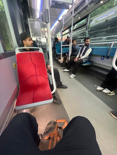 People are seated on a public bus. One individual is reclining on a red seat, while others are seated upright. The bus interior is visible, with windows showing the outside environment. A bag is placed on the floor next to a passenger in the foreground. The scene suggests a typical urban commute.