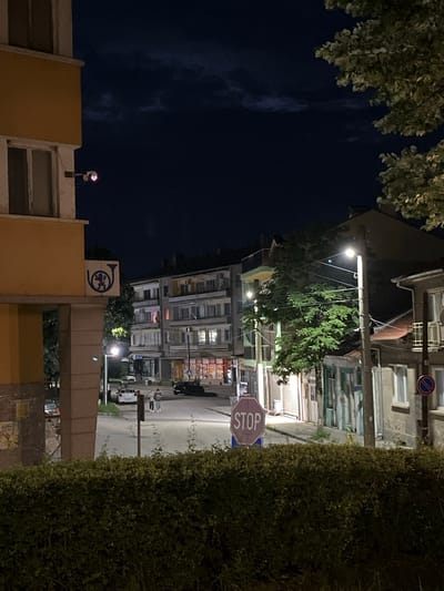 The image depicts a nighttime street scene in Veliki Preslav, Bulgaria, specifically at the intersection of ulitsa "Boris I" and ulitsa "Tsar Simeon Veliki," looking northeast from a slightly elevated vantage point.
The foreground is occupied by a dense, dark green hedge. To the left, a multi-story building, painted yellow with white trim, is partially visible. A security camera with a distinct red glowing LED is mounted on its facade. Below, affixed to a supporting pillar of this yellow building, is the blue logo of Bulgarian Post, featuring a postal horn with a lion's head. Multiple posters are visible on the lower section of the pillar, one of which contains the Cyrillic word "ТЕАТЪР" (Theatre).
The asphalt street is illuminated by several modern streetlights. Two individuals are walking on the sidewalk on the left side of the street, near a parked white sedan. A dark-colored vehicle is parked further down the street on the right. A prominent purple-red octagonal "STOP" sign stands on the right side of the street, partially obscured by the foreground hedge. Further along the street on the right, a blue circular sign with a red diagonal line (indicating no stopping or parking) is mounted on a building.
Across the street, a multi-story apartment building with numerous windows, some showing interior lights, is visible. To its right, a row of older, predominantly two-story buildings line the street. These include structures with closed shop fronts on the ground floor and one building with a distinctive light green facade. Trees with dense green foliage are distributed throughout the scene, particularly along the street and in the upper right, where a large tree branch with light-colored leaves extends into the frame. The nighttime sky is dark with visible cloud formations.