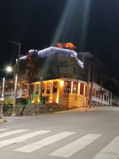 The image depicts a multi-story building illuminated at night in Villa Carlos Paz, Argentina. The building's facade features a stairway adorned with white lights and multiple sections with warm, yellow lighting. Green spotlights are positioned around small palm trees at the base of the building. A street with white crosswalk stripes is visible to the right of the building. A street light is on the lower left of the image. A sign spelling out "HOTEL" is visible near the bottom of the image.