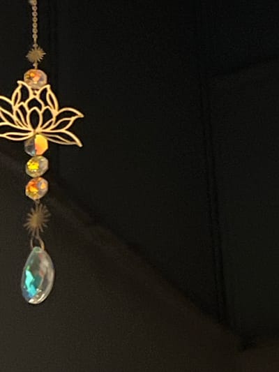 The image displays a close-up of a decorative object set against a dark, seemingly black, textured background. The primary subject is a metallic, golden-toned outline of a stylized flower, possibly a lotus, which forms a central part of a chain.

Attached to this flower outline is a delicate chain adorned with multiple faceted, iridescent beads. These beads are generally round or oval, and they reflect various colors such as yellow, orange, and hints of other spectrum shades. Further along the chain, smaller, star-shaped or spiky metallic elements are interspersed between the beads.

The chain culminates in a prominent, larger teardrop-shaped bead. This bead is highly reflective and iridescent, exhibiting distinct blue and green hues, suggesting it is made of crystal or glass.

There is no activity or event taking place; the image is a still photograph showcasing the details of this decorative item. The time of day, weather, and any emotions are not discernible from the image. No visible text is present. The dark background provides a strong contrast, allowing the golden metal and the shimmering, colorful beads to stand out prominently. The item could be a piece of jewelry or a charm.