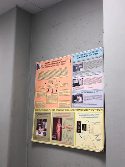 The image shows a poster on a light gray concrete wall. The poster is in Greek and shows a medical illustration. The poster is titled, "KATASKEUH FEELMEKENON ANATYIKON OPIANDON," meaning "Construction of Feelable Analytical Instruments." The poster is located at the coordinates 37.998, 23.82, in the city of Agia Paraskevi, Greece. The poster is attached to the wall with what appears to be tape. The poster is slightly askew. The image appears to be taken from a slightly elevated angle, with the camera pointed down at the poster.