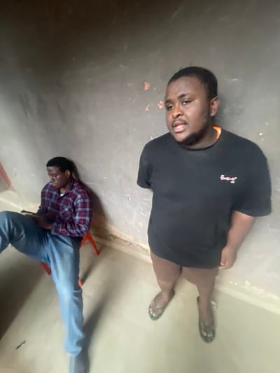 The image depicts two men indoors. One man, wearing a black t-shirt and flip-flops, is lying on the floor, looking up at the camera. The second man is also lying on the floor, wearing a red and blue checkered shirt and blue jeans. A red object, possibly part of a chair, is visible near the second man. The scene appears to be taking place in a room with concrete walls and floor.