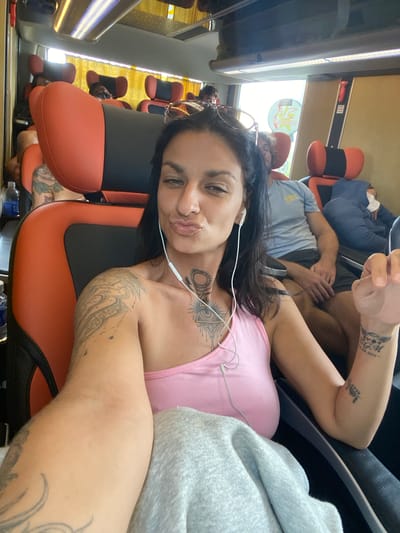 A woman with dark hair and tattoos on her arms and chest is taking a selfie on a bus. She is wearing a pink tank top and white earbuds. She is sitting in a red and black bus seat, and another person in a blue hoodie is sleeping in the seat behind her. The bus appears to be on a road in Đăng Srõn, Vietnam, as evidenced by the landscape visible through the windows and the yellow accents on the bus interior, consistent with local transportation aesthetics.