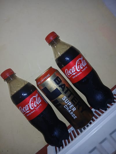 The image shows three drinks: two bottles of Coca-Cola and one can of Battery Super Charge. The Coca-Cola bottles are black with the iconic red label featuring the Coca-Cola logo and "ORIGINAL TASTE" text. One bottle is partially obscured, but both appear to be full or nearly full of a dark liquid and are covered in condensation, suggesting they are cold. The Battery Super Charge can is primarily gold and black, with "BATTERY SUPER CHARGE" prominently displayed in white text. It also has text that reads "+25% CAFFEINE". The items are arranged on a light-colored, textured surface, possibly a table or floor. In the background, a white and red striped grate or vent is visible, suggesting an indoor setting, possibly near a radiator or air vent. The lighting is somewhat dim, creating a slightly reflective surface on the bottles and can. The arrangement of the drinks is diagonal, with the can positioned between the two bottles. There are no people visible in the image. The scene is static and focuses solely on the beverages. The overall impression is that of a collection of drinks ready for consumption, possibly in a casual setting.