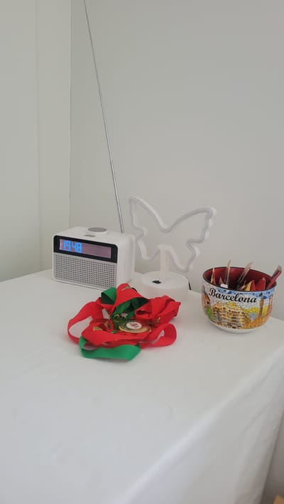A white table is covered with a white tablecloth. On the table are several objects: a white digital radio with a blue display showing "19:48", a metallic antenna extended upwards from the radio, a white neon butterfly-shaped light, a pile of red and green ribbons with two medals attached, and a decorative bowl filled with various small items. The bowl is colorful and has the word "Barcelona" printed on it, along with a mosaic-like depiction of buildings. The background is a plain white wall. There are no people visible in the image. The scene appears to be indoors, likely on a table. The city of Chippenham, United Kingdom is not depicted in this image.