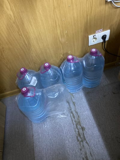 The image shows a wall with wood paneling and a white electrical outlet. A plug is inserted into the right socket of the outlet, and a black cord extends from it. A label on the wall above the outlet reads "220 B". In front of the wall, on the floor, are six large plastic bottles of water, mostly covered in plastic wrapping. The bottles have purple caps and appear to be partially full. The floor is covered in a textured grey material with some light brown stains. The setting appears to be indoors, possibly a storage area or a room in Luzhitsy, Russia.