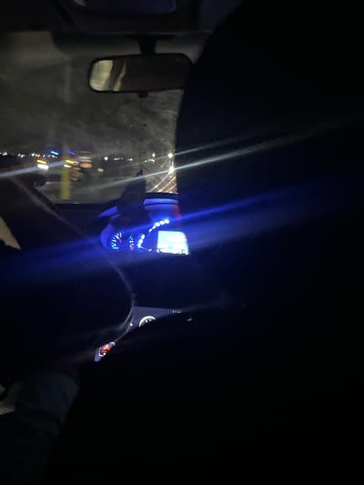 The image depicts the interior of a car at night, looking forward through the windshield. The dashboard is illuminated with blue and red lights, suggesting it is actively being driven. The view outside the car reveals a street scene with blurred lights of buildings and other vehicles, indicating movement through a city. In the foreground, a person's arm is visible, resting on the steering wheel. The overall impression is of a nighttime drive through a dimly lit urban area. The image does not provide enough visual information to determine the specific location within La Cruz del Pastel, Venezuela.