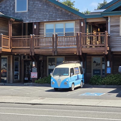 The image captures a front view of a blue and cream-colored Volkswagen Type 2 van parked in a lot in front of a wooden building in Cannon Beach, Oregon, United States. The building appears to be a commercial establishment, possibly a liquor store, with several windows and a covered walkway. A red "OPEN" sign is visible near the entrance. The van has a luggage rack on its roof and a sticker on its side depicting a coastal landscape. The parking lot has painted lines and a handicapped parking symbol. Behind the van, there are bushes and the building's facade. The sky is blue and clear.