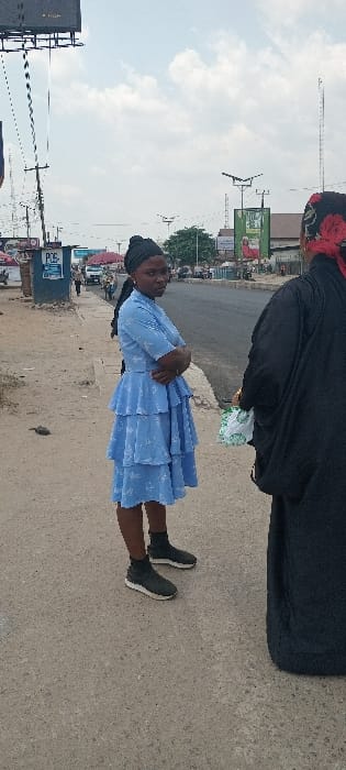 The image captures a daylight street scene in Aba, Abia, Nigeria, on an overcast day. Two individuals stand on a paved sidewalk in the foreground, adjacent to an asphalt road.
On the left, a young adult female with dark skin is visible. She wears a light blue, short-sleeved, tiered midi dress featuring subtle white patterns, black ankle-high sneakers with white soles, and a black headscarf. Her arms are crossed over her chest, and she stands facing slightly left, looking towards the viewer.
To her right, partially visible from the back, is another individual. This person wears a black, long-sleeved garment and a red and black floral patterned headscarf. A white plastic bag with green markings is held in their left hand.
The background shows an asphalt road extending into the distance, with a white van and other indistinct vehicles visible. Various commercial buildings, stalls, and street furniture line the road. A prominent blue structure displaying "POS" (Point of Sale) is situated behind the woman in blue. Multiple streetlights equipped with solar panels are present, along with large advertising billboards, including a dark one in the upper left and others with green backgrounds. Scattered trees are visible in the mid-ground.