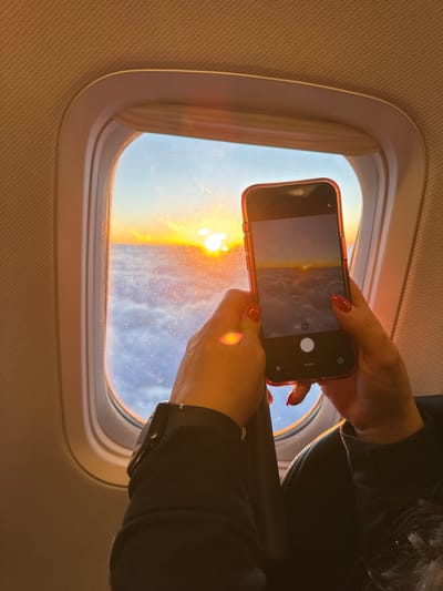 A person is taking a picture with their smartphone of the sunset outside an airplane window. The scene is lit by the warm hues of the setting sun. The phone is held by two hands with red nail polish. The airplane window is oval-shaped and dirty. The interior wall of the plane is textured. The photo was probably taken during a flight approaching Vecsés, Hungary, but there are no identifying elements that explicitly confirm it is Vecsés.