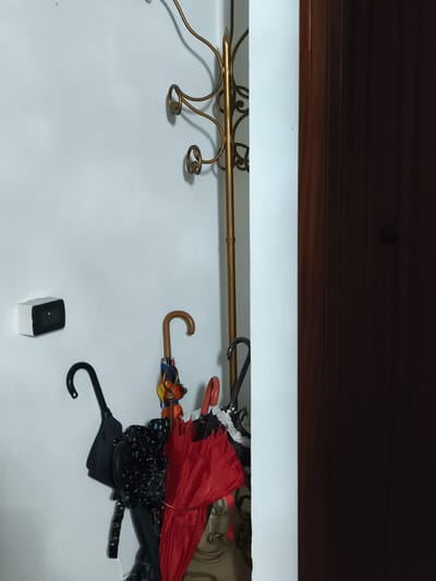 This media file captures a close-up, indoor scene. The primary subjects are a group of umbrellas hanging on a coat rack, next to a white wall and a dark brown door. The umbrellas display a variety of colors and patterns, including a bright red one, a black one with a white polka-dot design, and another with a multicolored, patterned fabric. One umbrella has a wooden handle.

To the left of the umbrellas, mounted on the white wall, is a small white device with a black frame and a red light illuminated in the center. This appears to be an electronic sensor or indicator.

The background consists of a plain white wall on the left and a portion of a dark brown wooden door on the right. A decorative, gold-colored coat rack with several hooks is positioned against the white wall, behind the umbrellas. The lighting suggests an indoor environment, possibly during the daytime with artificial light. There is no visible text in the image. The overall impression is one of an entryway or hallway where umbrellas are stored.