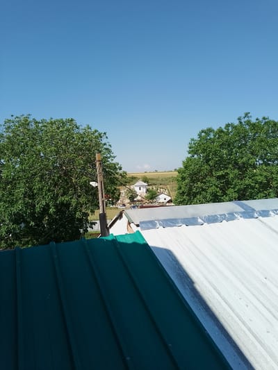 The image is a high-angle shot taken from a rooftop in Nicorești, Romania, on a clear, sunny day. The foreground is dominated by two sections of corrugated metal roofing, one dark green and the other light gray. Behind the rooftops, a lush green tree stands prominently on the left, with another on the right. In the middle ground, a village scene unfolds, featuring a white building with a pink roof, surrounded by gravestones, suggesting a cemetery. Further in the distance, rolling hills and fields are visible under a bright blue sky. A wooden utility pole with some wires is positioned between the foreground and the village.