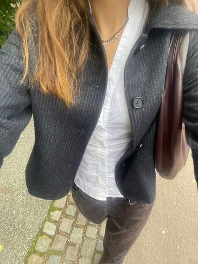 The image displays an individual in an outdoor urban setting in Fredensborg, Denmark, captured from a high-angle perspective focusing on the torso and legs. The subject wears a white collared shirt, unbuttoned at the neckline, underneath an unbuttoned dark grey pinstriped blazer with a single visible black button on the right lapel. A delicate silver-colored necklace featuring multiple small, reflective circular elements is visible around the neck. Dark brown trousers are worn below the waist. A dark brown leather shoulder bag is positioned on the individual's right side, partially visible. Long, light brown hair cascades over the left shoulder. The ground surface consists of a light grey asphalt or paved area on the left and a section of irregularly shaped cobblestones, exhibiting various grey and brown tones with some moss growth, extending across the right and lower portions of the frame. No specific actions or interactions with other subjects are apparent.