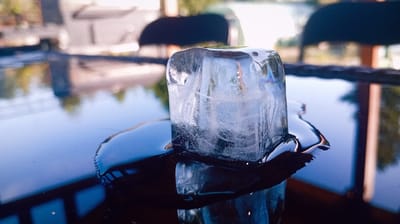 A single ice cube, partially melted, rests on a dark, reflective surface. The ice cube is transparent with some internal cloudy textures and small air bubbles, and its edges are softened by melting. A puddle of water surrounds the base of the ice cube, also reflecting the surroundings. The background is out of focus, showing blurred shapes of trees and possibly outdoor seating, suggesting an outdoor setting like a patio or deck. The lighting indicates it is daytime, with sunlight creating reflections and highlights on the ice and water. The overall mood is tranquil, perhaps suggesting a warm day and a refreshing element. No text is visible.