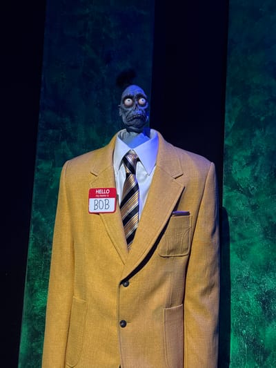 The image shows a headless mannequin wearing a yellow suit jacket, white collared shirt, and a striped tie. A name tag that reads "HELLO my name is BOB" is pinned to the jacket. The mannequin's head, which is a detailed, blueish-grey sculpt of a monstrous face with large, bulbous eyes and grimacing teeth, is positioned on top of the suit. The background is dark with hints of green and blue lighting, possibly simulating a stage or exhibit setting. There is no information in the image to suggest a location in Monterrey, Mexico.
