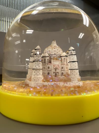 The image shows a close-up view of a snow globe with a model of the Taj Mahal inside. The Taj Mahal is made of white material and has black accents on its towers. The base of the snow globe is bright yellow. The interior of the globe contains an orange-colored material with iridescent glitter. The globe is sitting on a flat surface, likely a desk or shelf. In the background, out of focus, are vertical ribbed lines, possibly part of a filing cabinet or shelving unit. The scene is indoors, with overhead lighting reflecting on the curved glass surface of the snow globe. There is no indication of the city of Columbia, United States of America, or any specific location.