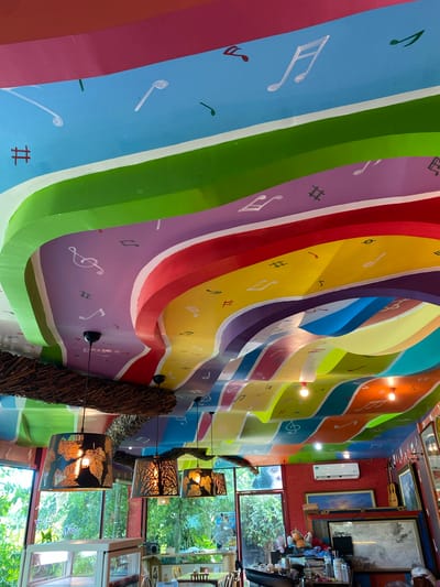 The image shows the interior of a brightly decorated restaurant or cafe in Bang Sare, Thailand, with a vibrant, multi-colored ceiling adorned with musical notes. The ceiling is curved and appears to be made of layered panels painted in stripes of pink, green, purple, red, yellow, blue, and orange. White musical notes, clefs, and hash symbols are painted on these colored panels. Hanging from the ceiling are three decorative lamps with cut-out designs of leaves and trees, illuminated from within. One lamp is larger and more prominent, made of what appears to be woven sticks. In the background, through large windows, lush green foliage is visible, suggesting an outdoor garden or natural surroundings. Inside, the walls are painted red brick and there are several framed artworks leaning against them. Electrical appliances, such as an air conditioning unit, are also visible. The overall impression is of a whimsical and artistic space.