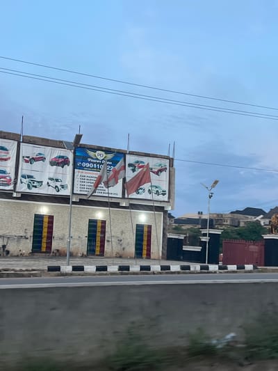 In Orifite, Nigeria, a medium-shot captures a building exterior under a twilight sky with scattered clouds and power lines. The building features a brick facade with prominent, brightly colored doors: red, yellow, and green. Above the doors, a large billboard displays various car models and advertisements for "Power Oven," listing services such as car painting, buffing, conditioning, and repair. Two flags are attached to poles in front of the billboard, one white with a red cross and another red flag. A modern street light stands near the building. To the right of the building, a dark-colored gate with pillars and a red metal fence is visible, with a lion statue positioned on the right side of the frame, partially obscured. A paved road with black and white markings runs in the foreground.