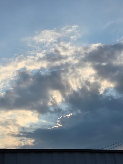 The image is a vertical, medium-shot photograph of a cloudy sky taken at an upward angle from a low vantage point. The sky is a mix of blue and grey clouds, with patches of sunlight breaking through. The clouds are dense and textured, with some appearing dark and heavy, while others are lighter and wispy, illuminated by the sun. In the lower portion of the frame, a dark grey, corrugated roofline is visible, suggesting the image was taken from near ground level, possibly looking up from behind a building. Two thin, dark wires stretch across the upper right corner of the frame, above the roofline. The overall impression is of a dramatic sky, possibly preceding or following a storm, with the sun fighting to penetrate the cloud cover. The location of Ермолаево, Russia is not discernible from the image content.