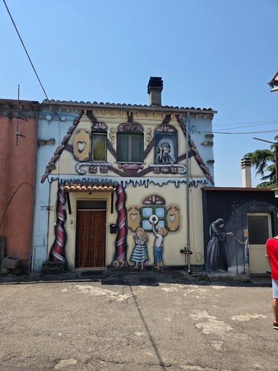The image shows a colorful, cartoon-like mural painted on the facade of a building in Viterbo, Italy. The mural depicts a whimsical scene, possibly inspired by a gingerbread house. Two children are shown playing with a soccer ball in front of the house, while another figure, dressed in a dark robe and hood, appears in a mural on an adjacent wall, interacting with a child. The building itself has a light blue and cream-colored exterior, with brown trim and a tiled roof. There are several windows and a prominent doorway covered by a curtain. The ground in front of the building is paved with asphalt, and there are some electrical wires visible against a clear blue sky. The overall impression is one of playful and imaginative street art.