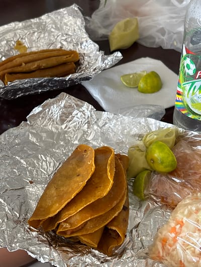 This image presents a close-up of food items typically associated with Mexican cuisine. Specifically, it features tacos, limes, cabbage slaw, and salsa packaged for takeout. The tacos appear to be arranged on a piece of aluminum foil, while the limes are both whole and sliced, some on a napkin, and some in a plastic bag with salsa. The slaw is packaged in a separate plastic bag. The scene appears to be indoors, on a dark brown surface. Given the items present, it is likely that this food was purchased from a taqueria in Monterrey, Mexico, where this type of food is common.