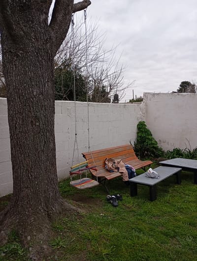 The image shows a backyard with a large tree on the left. A swing hangs from the tree, with a wooden seat decorated with colorful stripes. In the background, there is a white brick wall. A wooden bench with some clothing on it sits in front of the wall, and to the right are two dark gray tables. On the grass in front of the bench, a pair of black and white athletic shoes are placed. The ground is covered in green grass, and the sky is overcast. This scene is consistent with a backyard in the city of Ensenada, Argentina.