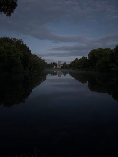 This media file depicts a surreal, almost mirrored landscape, likely created by reflection in water. The top half of the image shows a twilight sky with heavy, dark clouds. A line of trees with lush green foliage forms a distinct edge between the sky and the reflected scene.
The reflected scene, which occupies the bottom half of the image, mirrors the trees and sky. In the center of the reflection, a building with orange and red elements is visible, suggesting it might be a house or a small structure. The overall impression is one of a serene, possibly slightly melancholic, twilight atmosphere. The dark water reflects the cloudy sky and the trees with a high degree of clarity, creating a sense of depth and symmetry. No people are visible in the scene. The time of day appears to be dusk or early evening, given the dim lighting and the overcast sky. The location cues, such as the trees and the building, are typical of a rural or suburban setting. The overall mood is tranquil and contemplative. No visible text is present in the image.