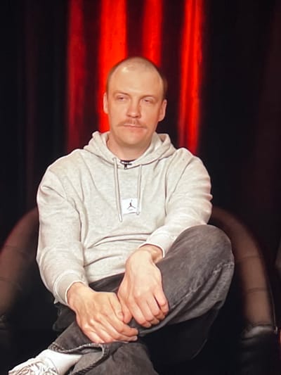 A medium shot captures a bald man with a mustache sitting in a dark chair. He is wearing a light grey hoodie with a small black logo on the left chest and dark grey jeans. His left leg is crossed over his right, with his hands resting on his lap. He is looking slightly to his left with a neutral expression. The background consists of dark curtains with vertical red lighting. The image appears to be a still from a video recording or photograph taken in an indoor studio setting, possibly in Genemuiden, Netherlands, given the context.