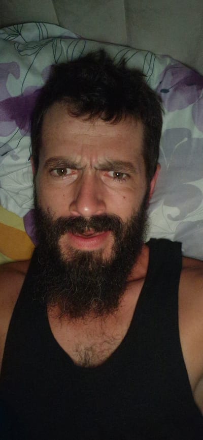 A man with dark, messy hair and a beard is lying down, looking up at the camera with a pained or disgusted expression. His eyebrows are furrowed, and his mouth is slightly open. He is wearing a black tank top. The background appears to be a bed with a patterned pillow. The image was captured in Wattenwil, Switzerland.